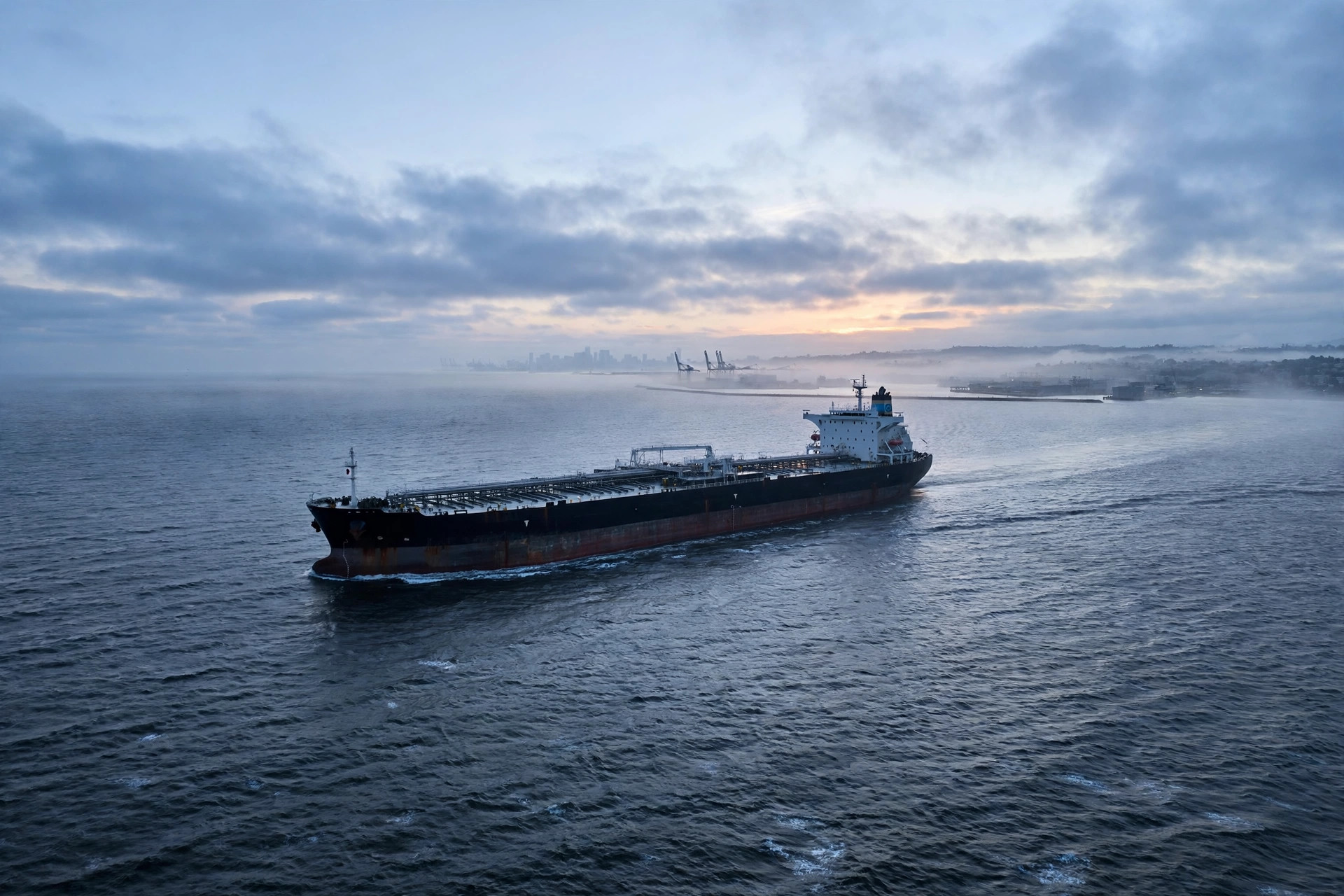 A large tanker ship sailing on open water toward a distant port at dawn