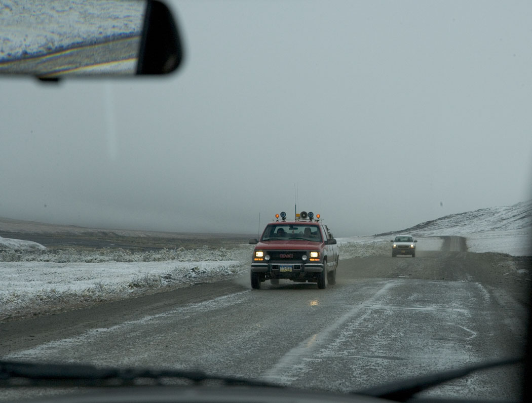 Two pickup trucks on a snow-dusted stretch of the Dalton Highway in Alaska under gray skies, seen through a windshield
