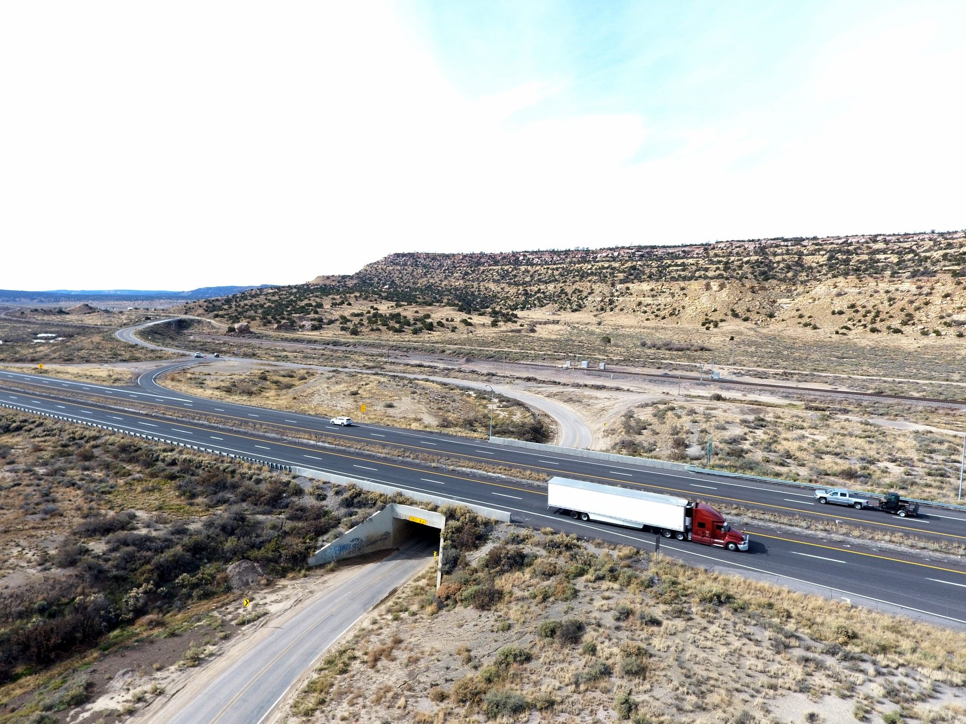Aerial view of a red semi truck traveling on Interstate 40 in western New Mexico, with the parallel rail line visible in the background