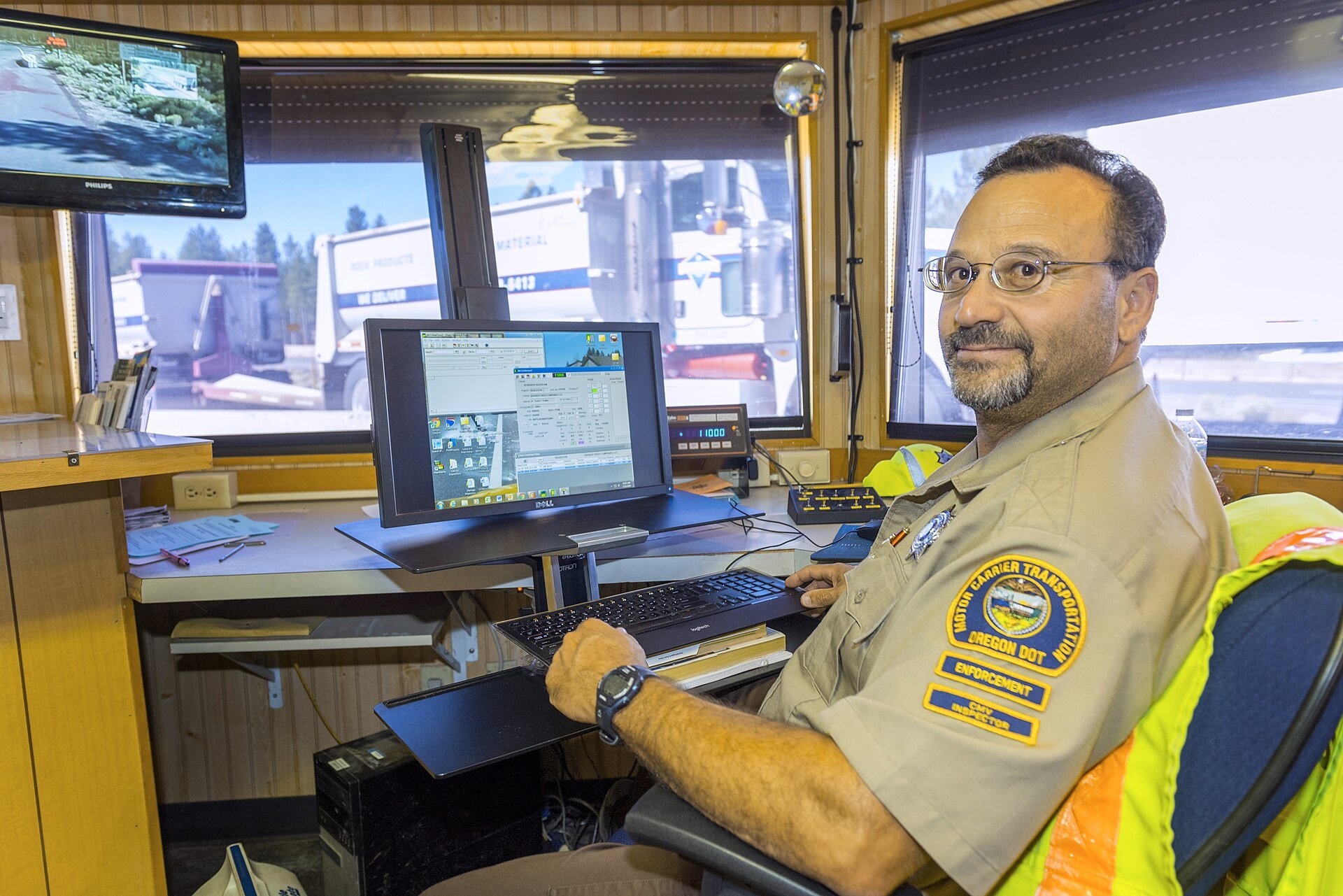 An Oregon DOT commercial vehicle inspector at his computer inside a weigh station, with semi trucks visible through the window behind him